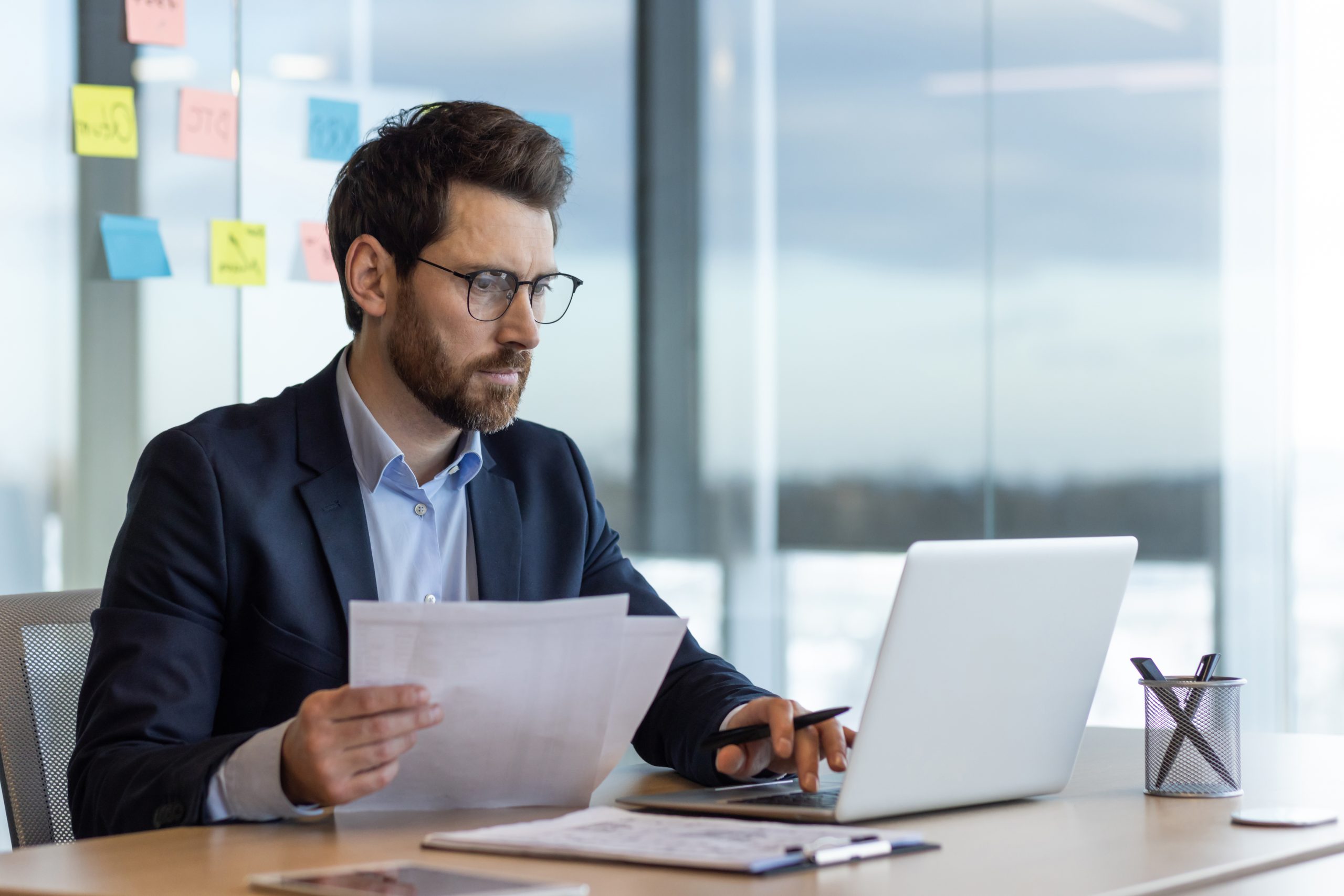 A professional man in a suit sits at a desk in a modern office, reviewing printed documents while working on a laptop, with sticky notes visible on a glass wall behind him.