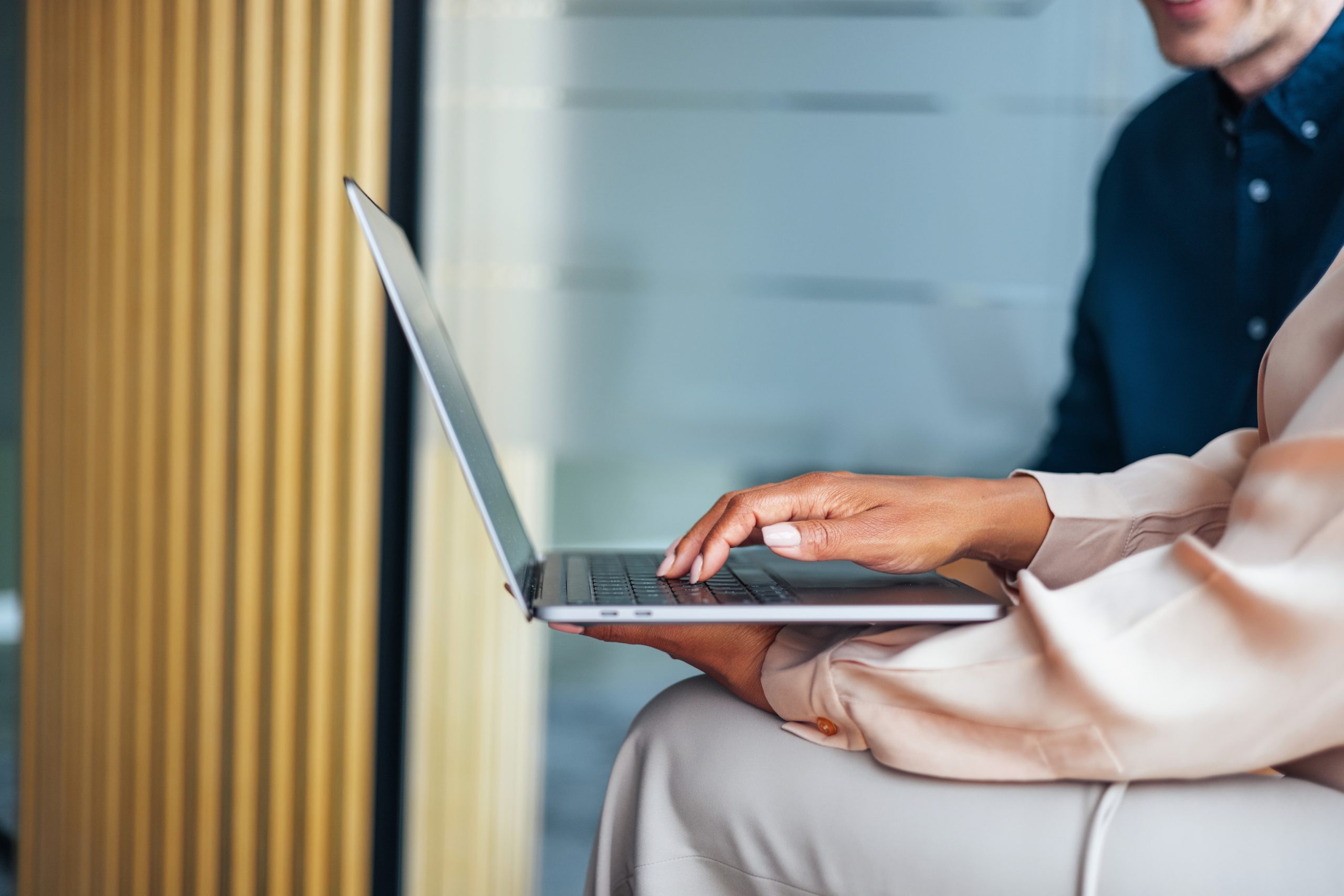 Close-up of a person typing on a laptop while seated, with another individual partially visible in the background in a modern office setting.